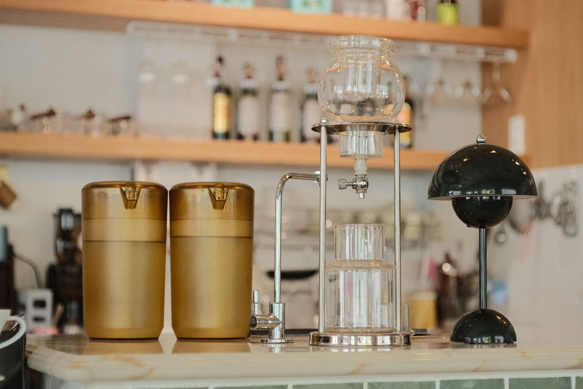 A brass bar cart with glass shelves holding a carafe, mugs and canisters