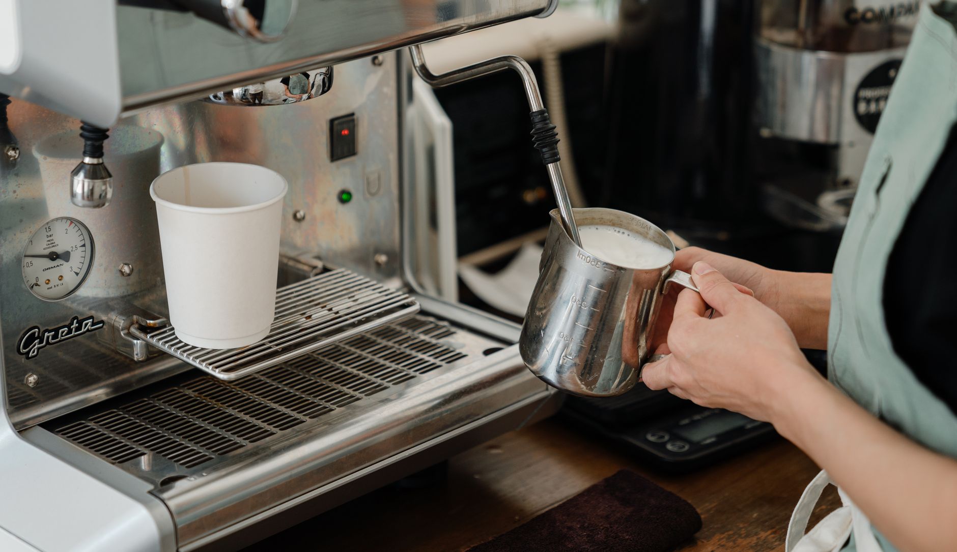 Stainless steel pitcher of steamed milk being poured into a wide ceramic cup